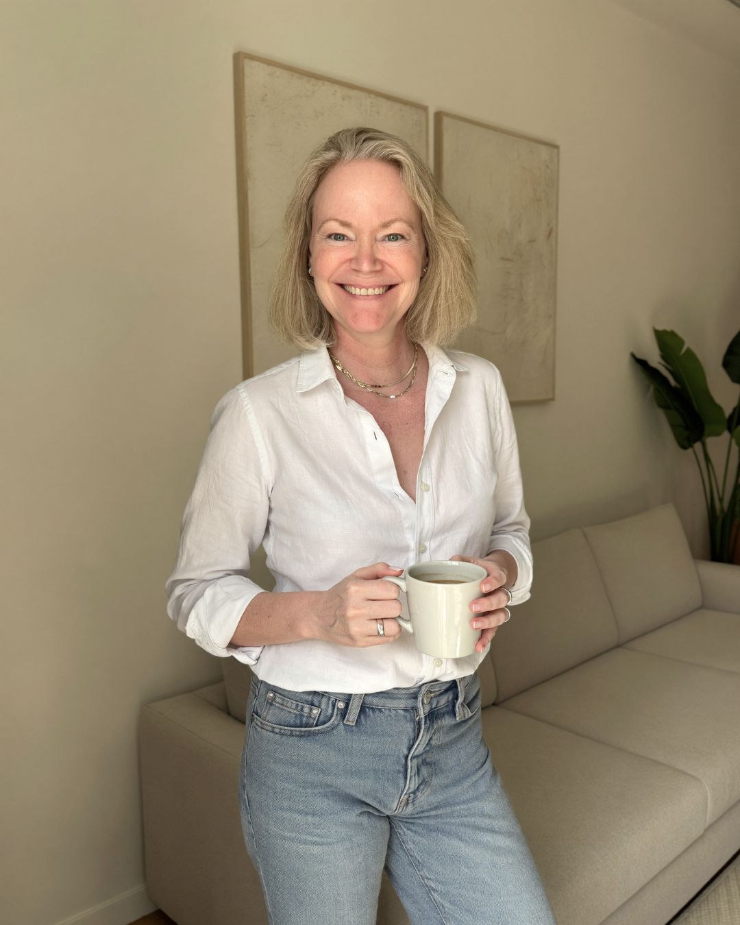 Nina Kolari seated on a stool in a white shirt and jeans, smiling against a black background